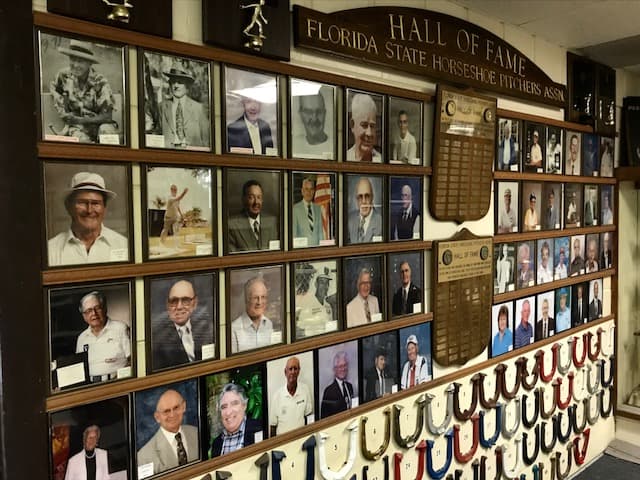 The Hall of Fame wall in the clubhouse, rows of inductee portraits and plaques above a case of championship horseshoes